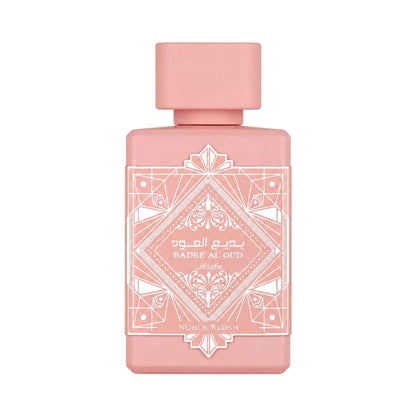 A rectangular pink perfume bottle with a matching pink cap, featuring white geometric and ornamental label detailing. The label reads 'Badee Al Qud' and 'Noble Blush'.